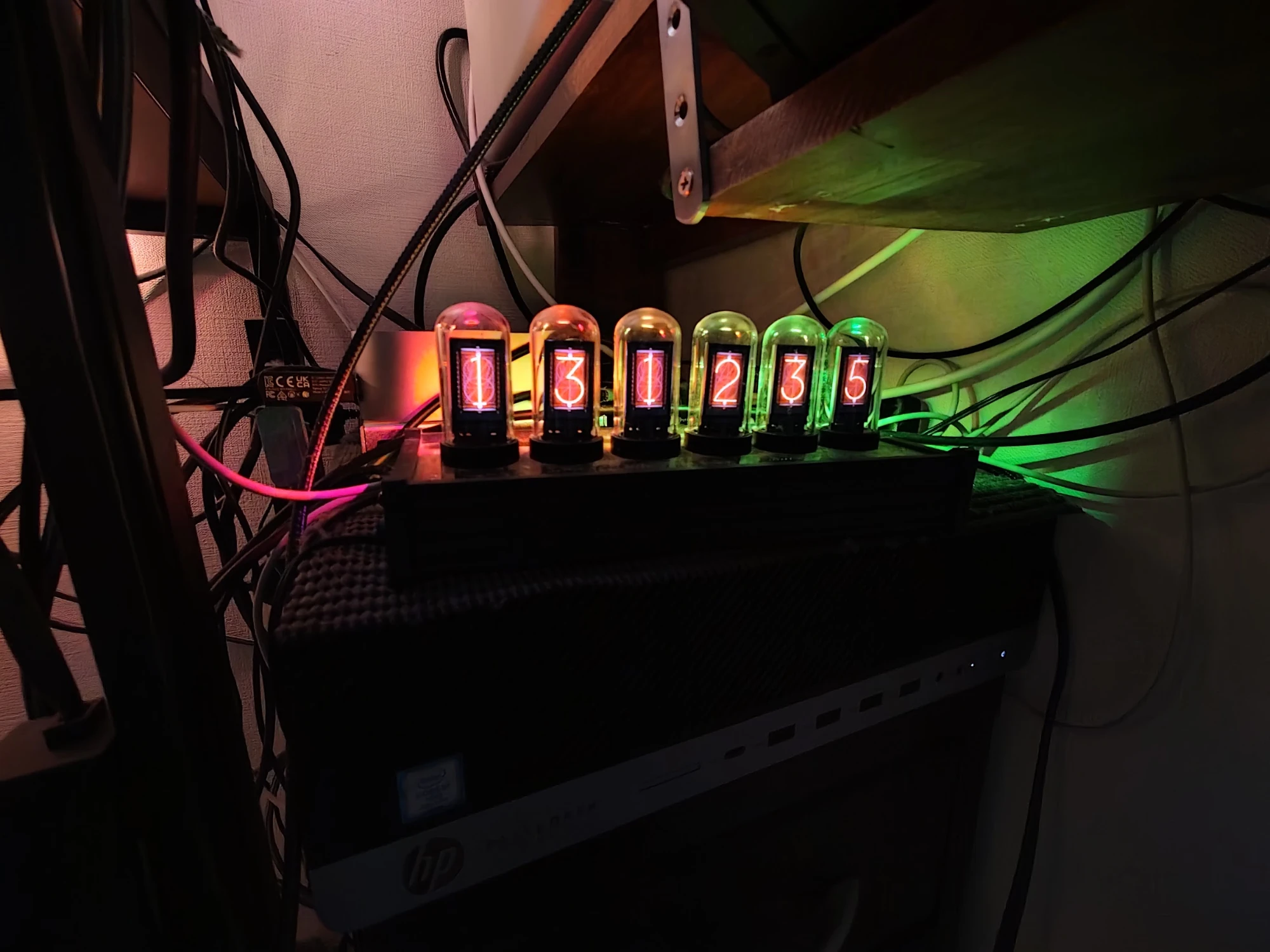 The EleksTube IPS clock in a dark corner of the room, under a wooden shelf and tangled cables: six glass tubes glowing orange-red with nixie-style digits reading 13 12 35, lit from behind by pink, yellow and green backlight LEDs.
